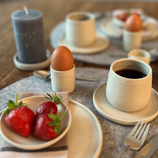 zwei beigefarbene handgedrehte Tassen aus Steinzeugton mit passender Untertasse auf einem Frühstückstisch