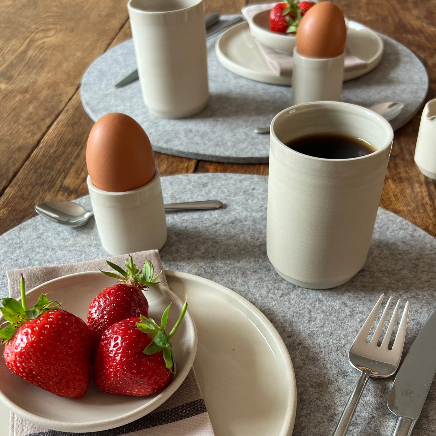 zwei beigefarbene handgedrehte Becher aus Steinzeugton mit Kaffee gefüllt auf einem gedeckten Frühstückstisch