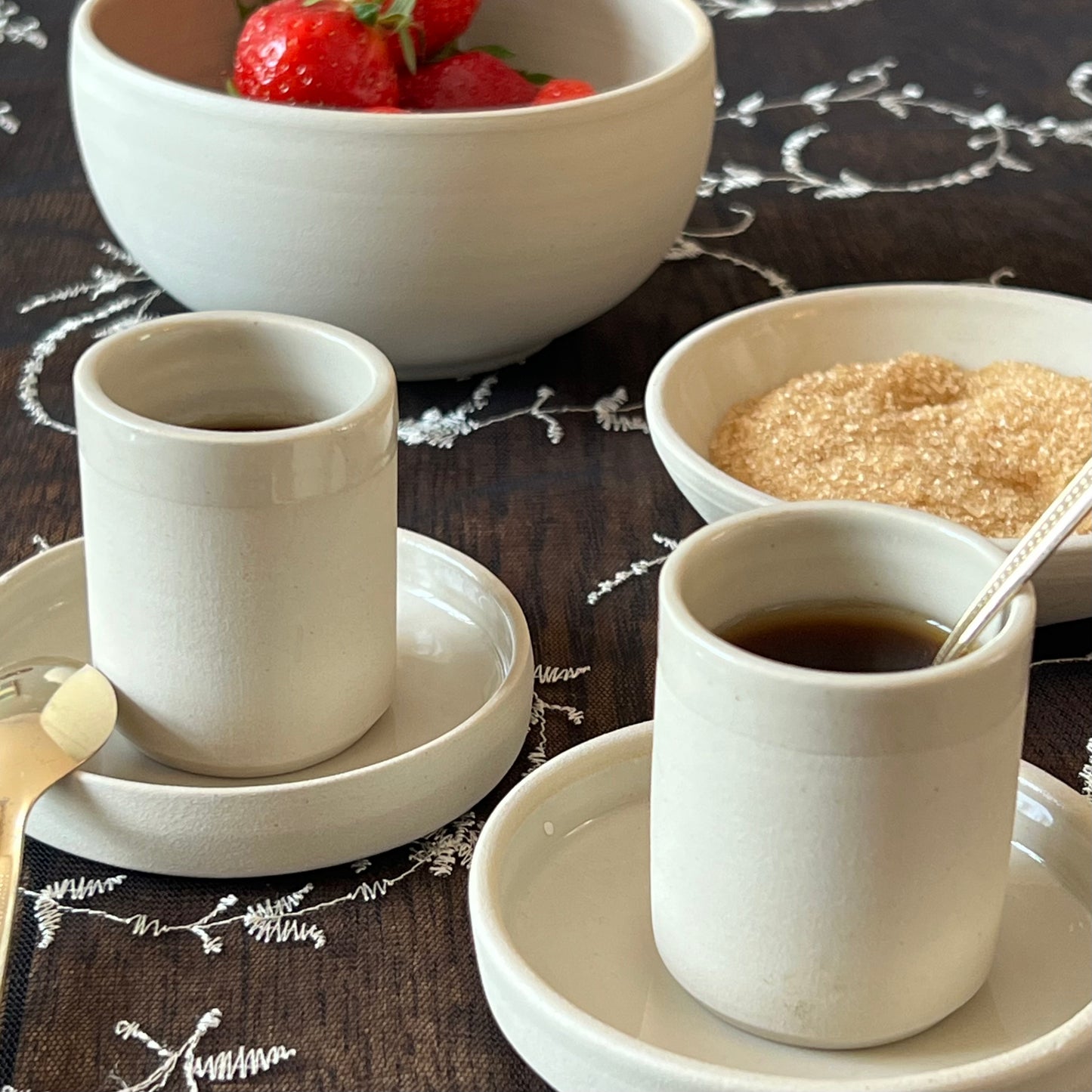 zwei beigefarbene handgedrehte Espressotassen aus Steinzeugton auf passenden Untertassen neben Zucker- und Obstschälchen