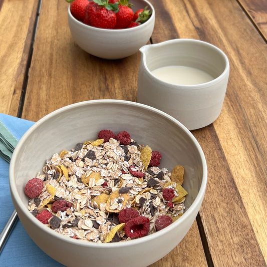 große beigefarbene handgedrehte Bowl aus Steinzeugton mit Müsli gefüllt, daneben Milch und Erdbeeren
