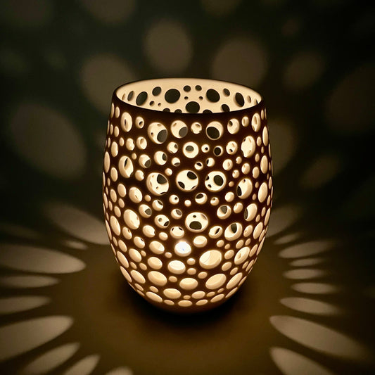 im Dunkeln aufgenommenes Porzellanlicht mit vielen sehr eng liegenden unterschiedlich großen Löchern aus dem eine Kerze ein sehr gemütliches Licht wirft