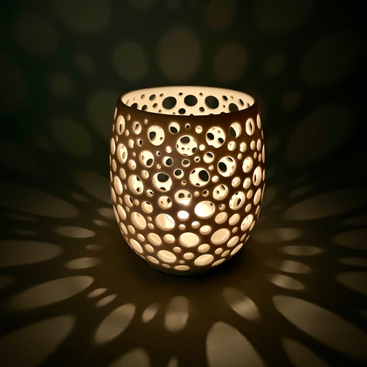 im Dunkeln aufgenommenes Porzellanlicht mit vielen sehr eng liegenden unterschiedlich großen Löchern aus dem eine Kerze ein sehr gemütliches Licht wirft