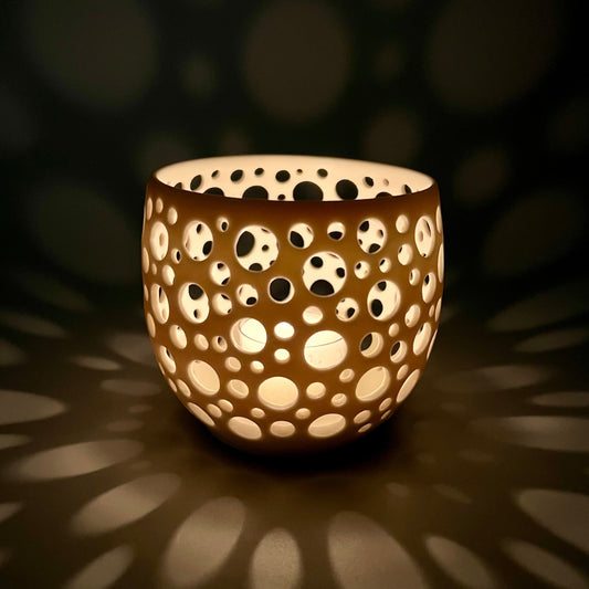 im Dunkeln aufgenommenes Porzellanlicht mit vielen sehr eng liegenden unterschiedlich großen Löchern aus dem eine Kerze ein sehr gemütliches Licht wirft