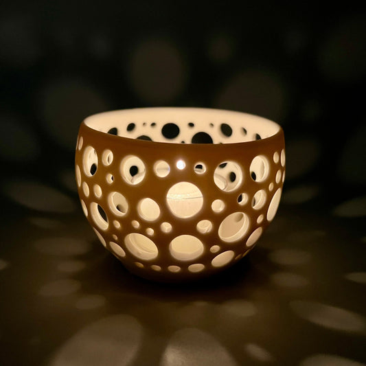 im Dunkeln aufgenommenes Porzellanlicht mit vielen sehr eng liegenden unterschiedlich großen Löchern aus dem eine Kerze ein sehr gemütliches Licht wirft