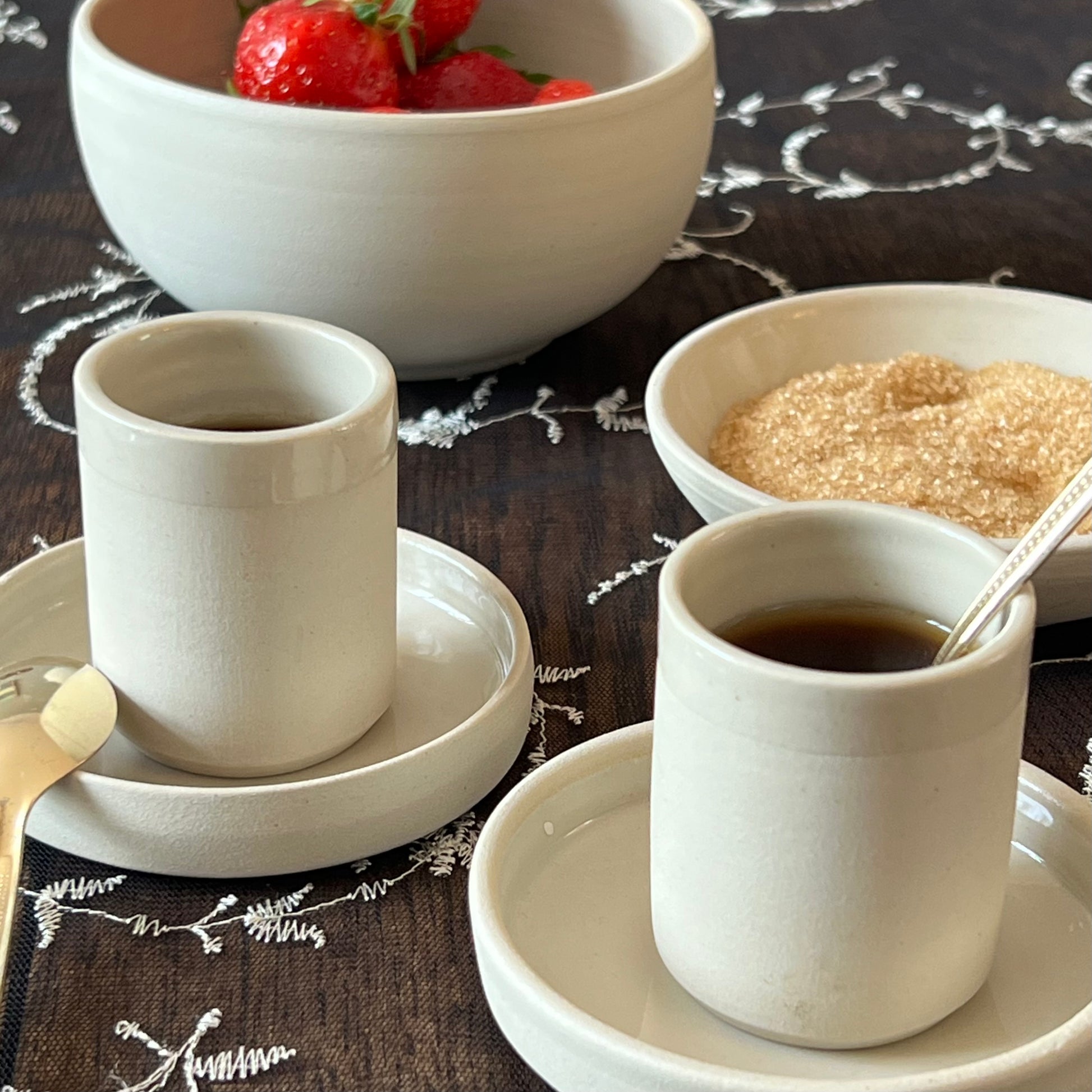 zwei beigefarbene handgedrehte Espressotassen aus Steinzeugton auf passenden Untertassen neben Zucker- und Obstschälchen