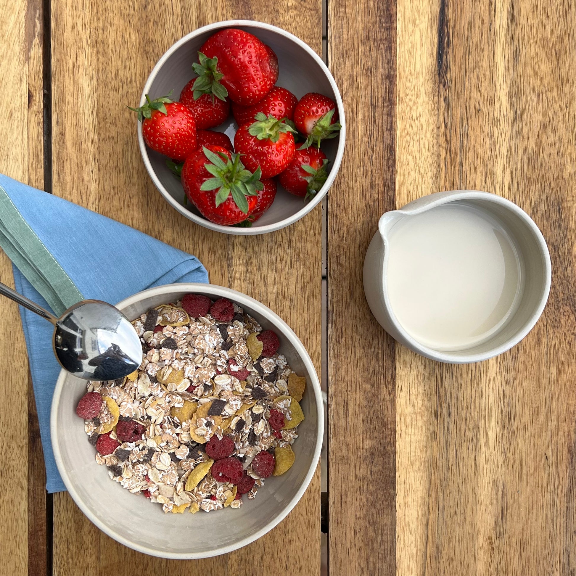große beigefarbene handgedrehte Bowl aus Steinzeugton mit Müsli gefüllt, daneben Milch und Erdbeeren