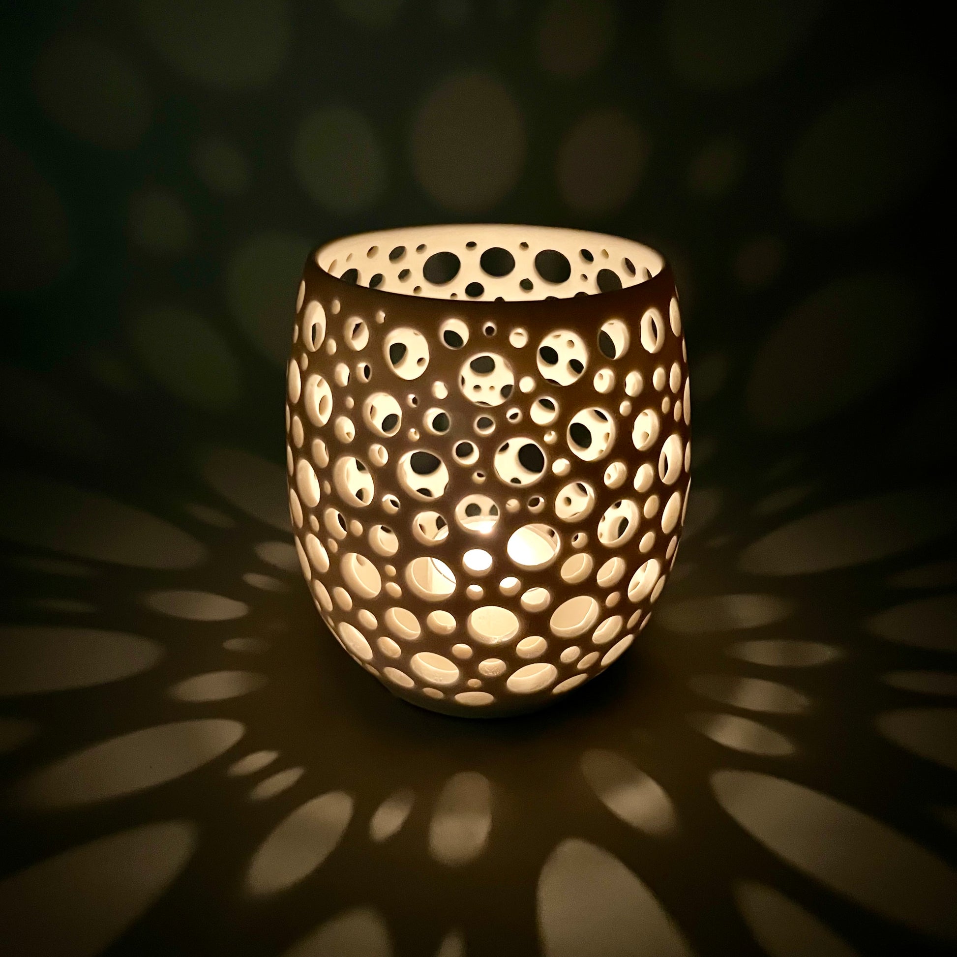 im Dunkeln aufgenommenes Porzellanlicht mit vielen sehr eng liegenden unterschiedlich großen Löchern aus dem eine Kerze ein sehr gemütliches Licht wirft