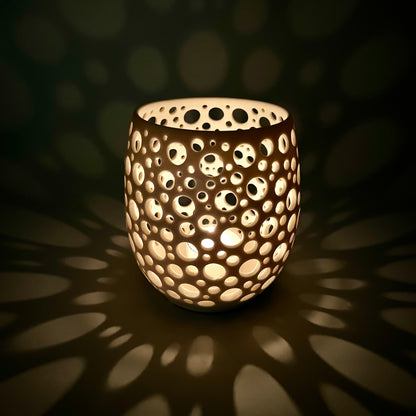 im Dunkeln aufgenommenes Porzellanlicht mit vielen sehr eng liegenden unterschiedlich großen Löchern aus dem eine Kerze ein sehr gemütliches Licht wirft